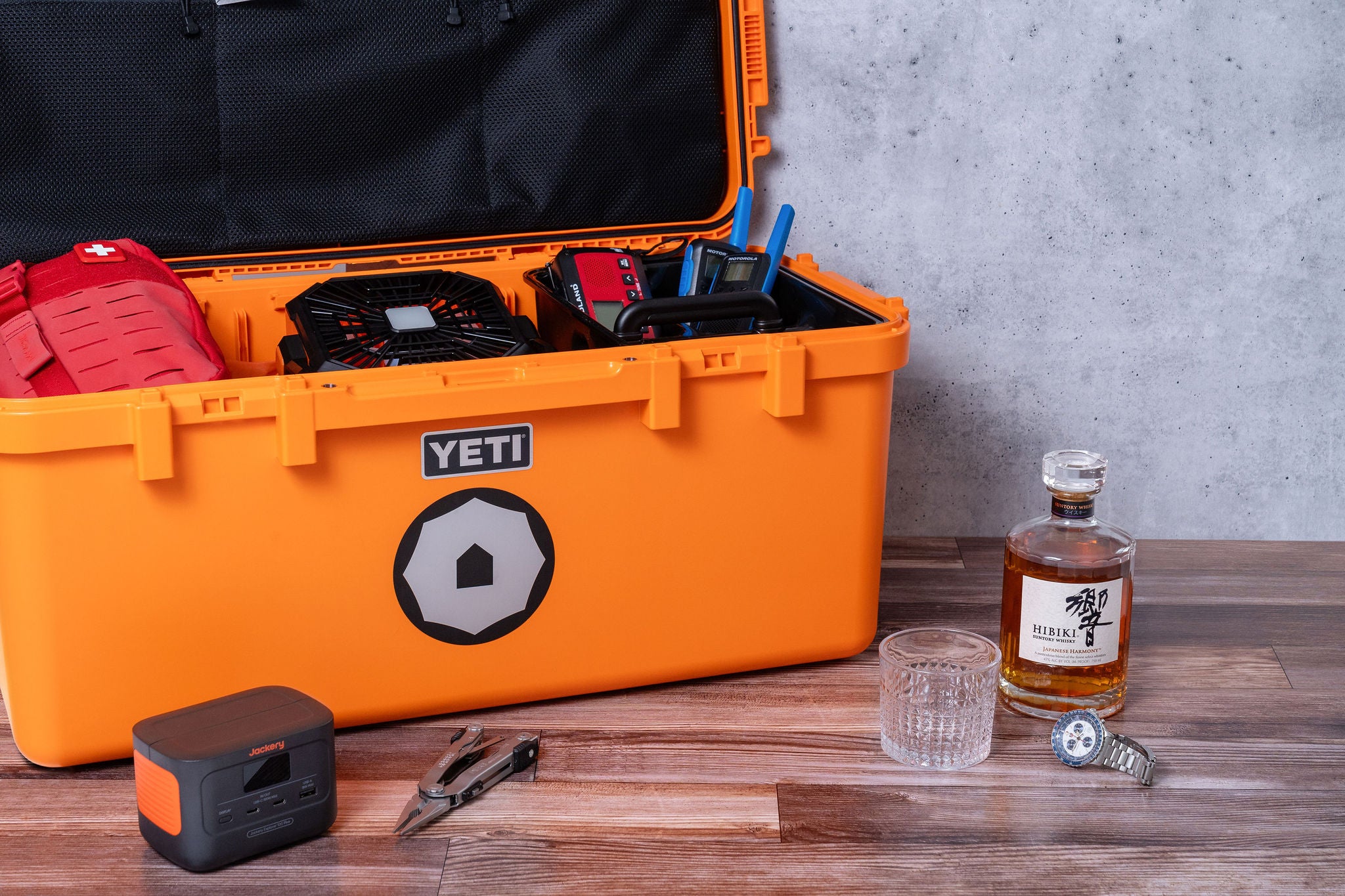 An open orange emergency kit box with various supplies arranged inside, including a bottle, glasses, and other emergency items.